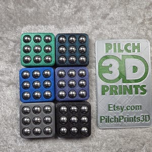 Könnte beinhalten: Sechs farbige, quadratische Fidget-Spielzeuge mit eingebetteten Metallkugellagern. Die Farben sind grün, dunkelgrün, blau, lila, grau und schwarz. Ein rechteckiges Schild mit dem Text "PILCH 3D PRINTS" und der Etsy.com-Adresse ist ebenfalls vorhanden.