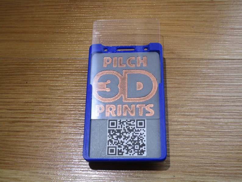 May include: A blue badge holder with a clear plastic cover. The badge insert features the text "PILCH 3D PRINTS" in copper-colored letters, along with a QR code. The holder is on a wooden surface.