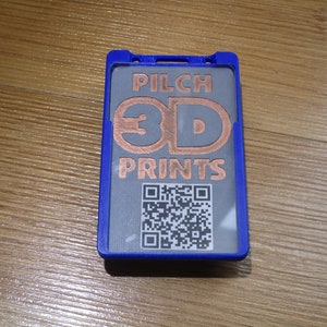 May include: A blue badge holder with a gray insert featuring the text "PILCH 3D PRINTS" in copper-colored letters. A QR code is printed below the text. The holder is on a wooden surface.