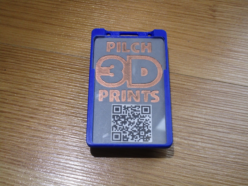 May include: A blue badge holder with a gray insert featuring the text "PILCH 3D PRINTS" in copper-colored letters. A QR code is printed below the text. The holder is on a wooden surface.