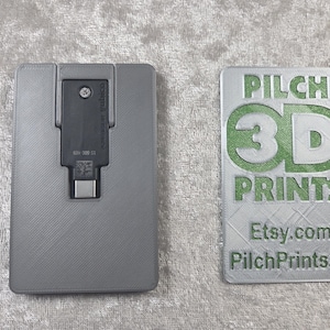Könnte beinhalten: Ein grauer, rechteckiger 3D-gedruckter Halter mit einem schwarzen Yubico-Sicherheitsschlüssel mit USB-C-Anschluss, neben einem grauen, rechteckigen 3D-gedruckten Schild mit grünem Text, der PILCH 3D PRINTS, Etsy.com und PilchPrints3D lautet.