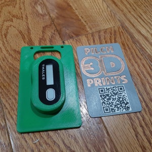 May include: A green 3D printed card holder with a black Thales security token inside. The card holder has a slot for a card and a hole for a lanyard. The card holder is next to a gray card with the text "PILCH 3D PRINTS" and a QR code.