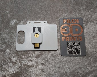 Pode incluir: Um porta-cartões de plástico branco com uma unidade USB cinza inserida. O porta-cartões tem uma ranhura para um cordão. A unidade USB tem uma letra dourada "Y" nela. O porta-cartões está ao lado de um cartão preto com o texto "PILCH 3D PRINTS" e um código QR.