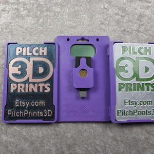 May include: A purple 3D-printed case with a USB-C connector and two hinged panels. Each panel features the text "PILCH 3D PRINTS" and "Etsy.com PilchPrints3D" in a contrasting color. The case is designed to hold a small electronic device.