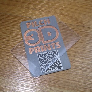 May include: A gray business card with the text "PILCH 3D PRINTS" in copper-colored lettering, along with a QR code. A clear plastic overlay is on top of the card. The card is on a wooden surface.