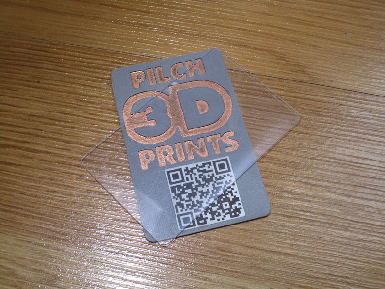 May include: A gray business card with the text "PILCH 3D PRINTS" in copper-colored lettering, along with a QR code. A clear plastic overlay is on top of the card. The card is on a wooden surface.