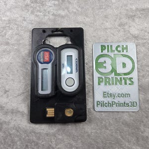 Può includere: Portachiavi di sicurezza nero stampato in 3D con due dispositivi di autenticazione digitale e un connettore USB. È presente anche un biglietto da visita bianco e verde con il testo "PILCH 3D PRINTS" e l'indirizzo Etsy.com.