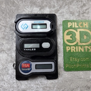 Può includere: Supporto nero stampato in 3D con tre token di sicurezza: HYPEROTP, THALES e RSA SecurID. Una carta verde con "PILCH 3D PRINTS" e l'indirizzo Etsy.com è accanto. Il supporto è progettato per contenere in modo sicuro i token.