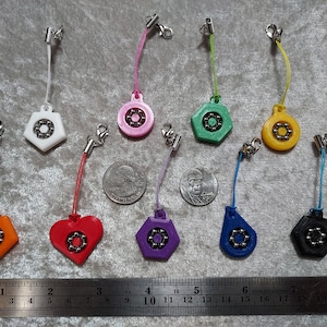 Puede incluir: Una colección de diez dijes de plástico de colores con rodamientos de bolas de metal en el centro, cada uno unido a un pequeño cordón con un cierre de langosta. Los dijes son blancos, rosas, verdes, amarillos, naranjas, rojos, morados, azules y negros, mostrados con una regla.