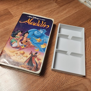 May include: A VHS tape case with a colorful image of the Disney movie Aladdin. The case is white with a blue and gold border. The title "A Walt Disney Classic Aladdin" is printed on the front of the case. The image features Aladdin, Jasmine, and Genie.