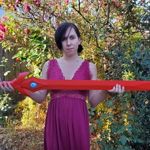 May include: A person holding a large, red, plastic sword with a blue gem in the center. The sword has a decorative design on the hilt.