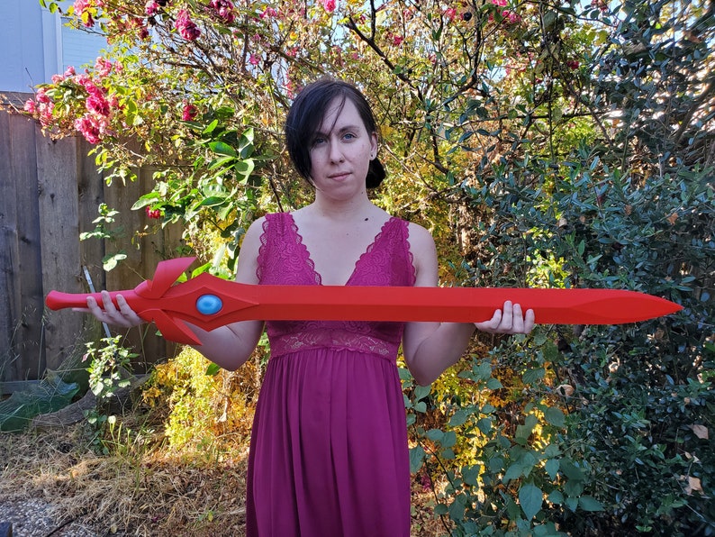 May include: A person holding a large, red, plastic sword with a blue gem in the center. The sword has a decorative design on the hilt.