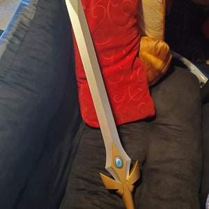 May include: A silver and gold sword with a blue gem in the center of the hilt. The sword is lying on a black couch with a red pillow behind it.
