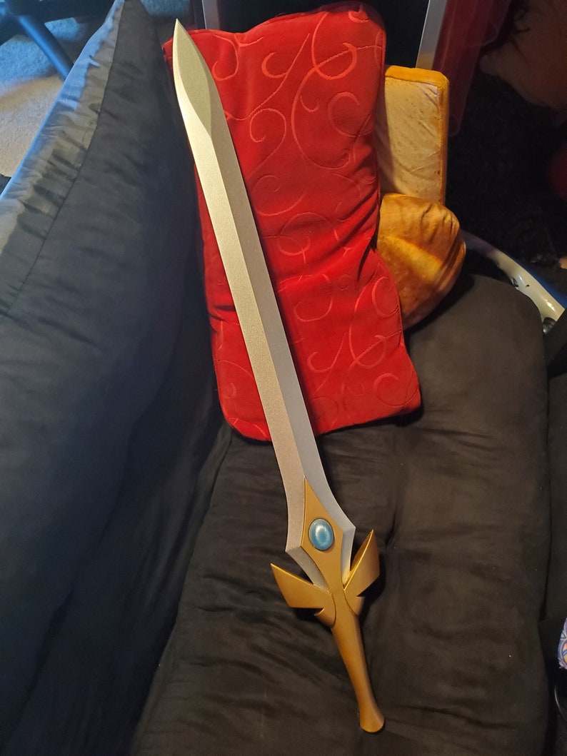 May include: A silver and gold sword with a blue gem in the center of the hilt. The sword is lying on a black couch with a red pillow behind it.