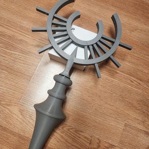 Peut inclure: Un bâton décoratif gris avec un motif de soleil stylisé en haut. Le bâton présente une poignée détaillée avec une forme bulbeuse. L'article est fait d'un matériau solide et repose sur une surface en bois.