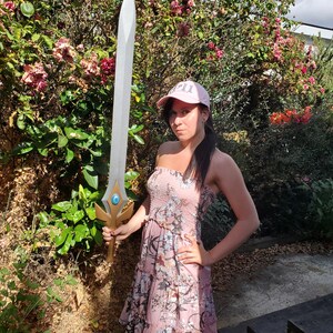 May include: A woman wearing a pink floral dress and a pink baseball cap with the word "GIRL" on it, holding a silver sword with a blue gem in the center.