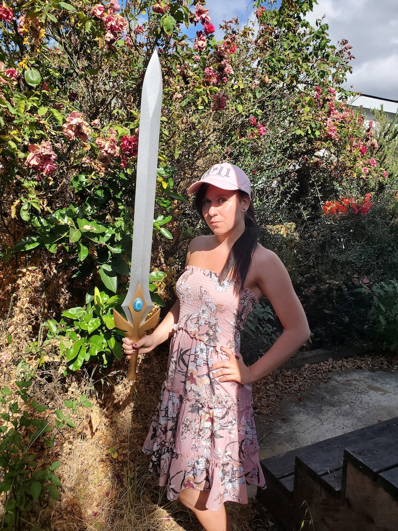 May include: A woman wearing a pink floral dress and a pink baseball cap with the word "GIRL" on it, holding a silver sword with a blue gem in the center.