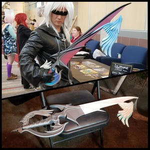 May include: A cosplay prop sword with a black, red, and blue blade, held by a person in a black jacket. The sword has a detailed hilt and a wing-like design. A second sword is displayed on a chair.
