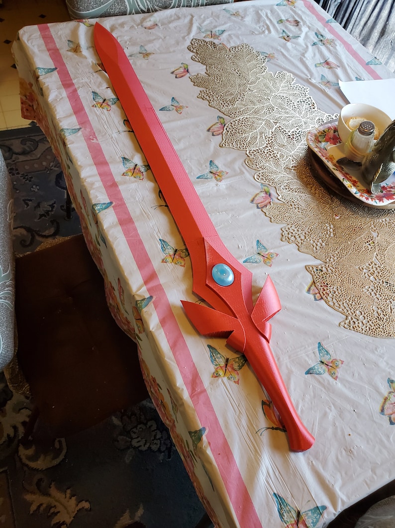 May include: A red and blue plastic sword with a decorative design. The sword is lying on a table with a floral tablecloth.