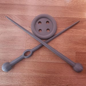 May include: A gray button with four holes and two gray knitting needles crossed over it. The needles have rounded ends and a loop on one side. The items are arranged on a wooden surface.