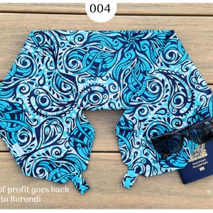 May include: A blue and white patterned travel neck pillow with sunglasses and a passport. The pillow has a repeating design of swirls and floral shapes. The text "004" is in the top center. The text "100% of profit goes back to Burundi" is at the bottom.