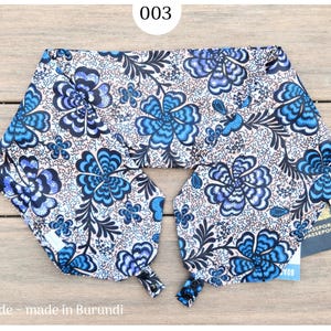 May include: A blue and white floral patterned travel neck pillow. The pillow has a repeating pattern of large blue flowers and smaller blue and black floral accents. The text "003" is in a white circle at the top. The text "fairtrade - made in Burundi" is at the bottom.