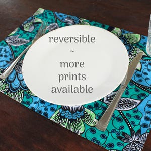 May include: A rectangular placemat featuring a vibrant teal, blue, and black floral pattern. A white plate sits in the center with the text 'reversible ~ more prints available' printed on it, accompanied by a silver fork, knife, and glass.