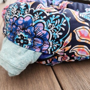 May include: Close-up of a pet bed with a vibrant floral pattern in blue, purple, and orange on a navy background. A light blue fabric is visible on the side. The bed is on a wooden surface.