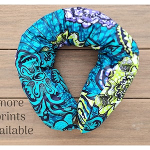 May include: A patterned neck pillow in teal, blue, and purple, featuring a floral design. The text "more prints available" is visible. The pillow is displayed on a wooden surface.