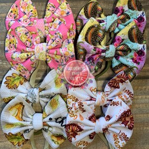 May include: Four fabric hair bows with elastic headbands. The top left bow features a pink background with colorful food illustrations. The top right bow has a yellow and blue floral pattern. The bottom left bow shows autumn leaves and food. The bottom right bow is white with turkey graphics and the text "GOBBLE TIL YOU WOBBLE!"