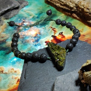 May include: A green gemstone pendant with a triangular shape hangs from a black lava rock beaded bracelet. The bracelet has a green cord and gold accents. The pendant and bracelet are on a dark gray surface.