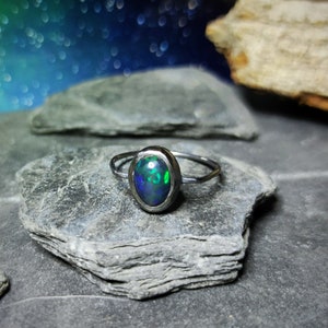 Puede incluir: Un anillo de plata con una piedra de ópalo de color verde oscuro y azul. El anillo está colocado sobre un fondo de piedra gris.