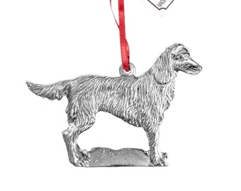 Op de afbeelding: Een zilveren tinnen ornament van een staande golden retriever hond. Het ornament heeft een rood lint om te hangen aan een kerstboom. Het ornament is gemaakt door House of M Pewter en heeft de tekst "Made in North Carolina since 1980".
