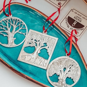 May include: Three silver tree of life ornaments on a teal blue plate with a red ribbon attached to each. The ornaments are in the shape of a circle, square, and rectangle. The plate has a brown rim. The text on the tag reads "House of Morzan Pewter Made in North Carolina Since 1981."