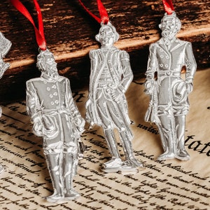 May include: Four silver ornaments depicting historical figures, each with a red ribbon attached. The figures are dressed in colonial-era clothing and appear to be standing on a piece of paper with text.