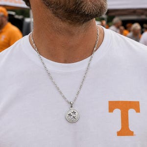 May include: A silver circular pendant necklace featuring three stars inside a circle, worn with a white t-shirt displaying an orange letter T logo, a white and orange baseball cap, and black sunglasses, reflecting a bright outdoor setting.