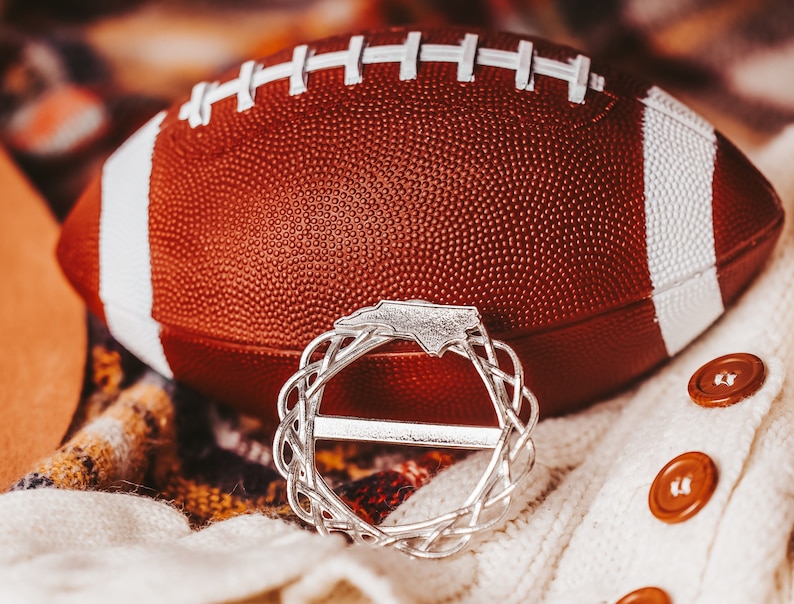 May include: A brown leather football with white stripes sits on a white and brown patterned fabric. A silver ring with a North Carolina state outline is in the foreground.