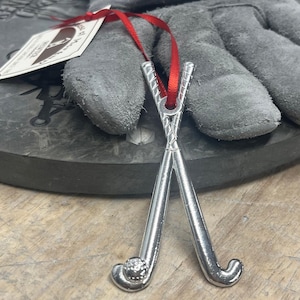 May include: A silver-colored hockey stick ornament with a red ribbon for hanging. The ornament features two crossed hockey sticks and a hockey ball. The ornament is likely made of metal and is designed to be a decorative item.