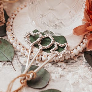 Op de afbeelding: Drie zilverkleurige toverstokken met verschillende vormen aan de toppen: een hart, een cirkel en een ster. De toverstokken liggen op een wit oppervlak met witte kralen en groene bladeren.