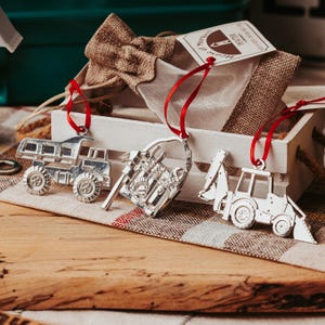 May include: Three silver metal Christmas ornaments shaped like a dump truck, a toolbox, and a backhoe. The ornaments are hanging from red ribbon and are displayed in a white wooden crate.