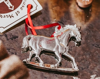 May include: A silver rocking horse ornament with a red ribbon. The ornament is on a dark brown surface. The ornament has the text "House of More" on it.