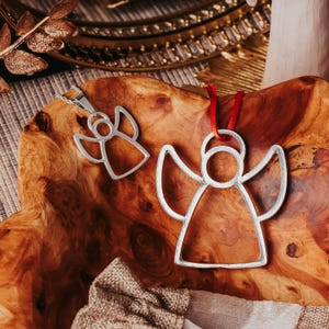May include: Two silver angel ornaments, one a pendant on a chain, the other a hanging ornament with a red ribbon. Both have a simple, open design. They rest on a wooden surface with a warm, reddish-brown tone, suggesting a holiday theme.