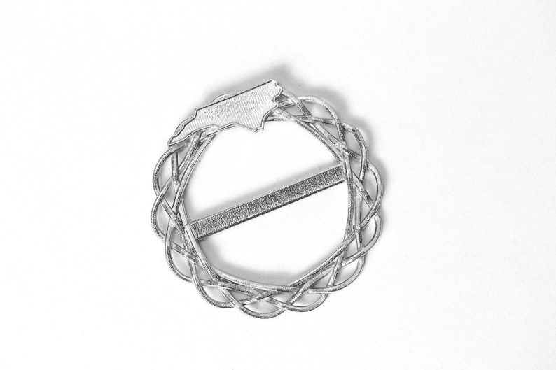 May include: Silver-toned metal belt buckle featuring a raised outline of the state of North Carolina. The buckle is circular with a woven design and a straight bar across the center.