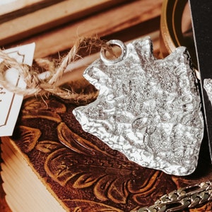 May include: Silver arrowhead pendant with a textured surface. The pendant is hanging from a piece of twine on a brown leather-bound book.