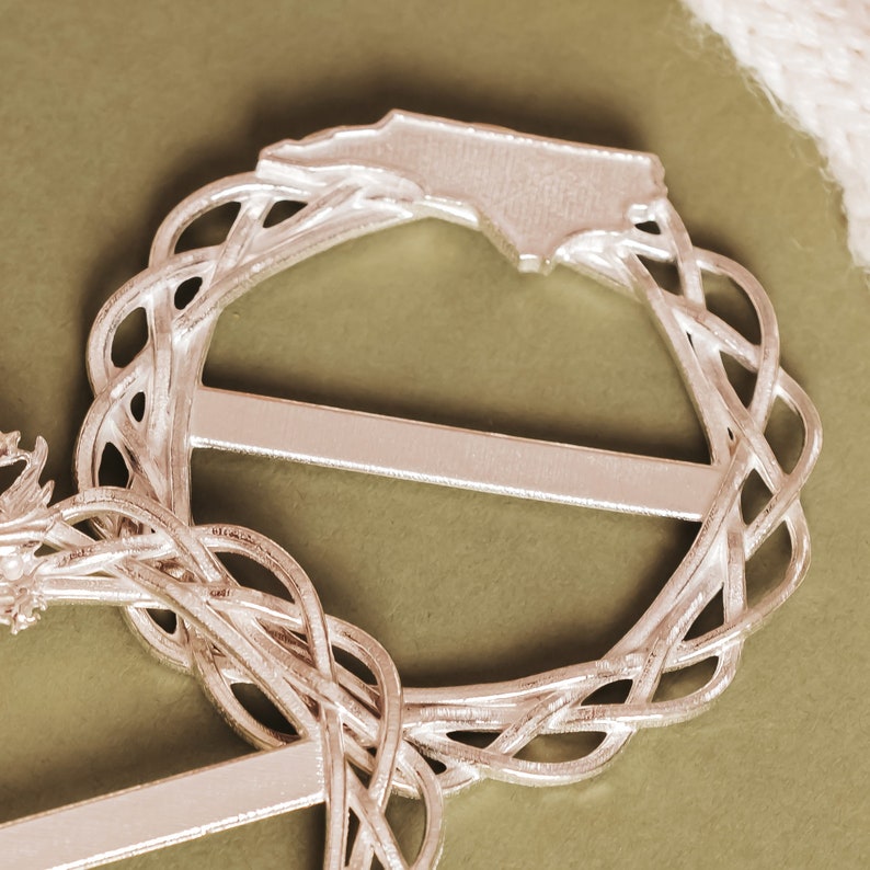 May include: Silver-toned metal belt buckle with a decorative design resembling a wreath. The buckle features a raised outline of the state of North Carolina.