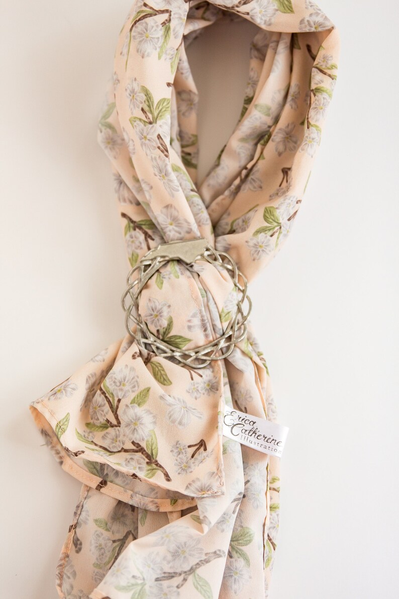 May include: A peach-colored silk scarf with a floral print of white flowers and green leaves. The scarf is tied with a silver metal ring with a braided design. The scarf has a white tag with the text "Erica Catherine Illustration" on it.