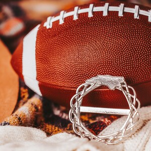 May include: A brown and white football with a silver charm in the shape of the state of North Carolina. The charm is a wreath with the state outline in the center.