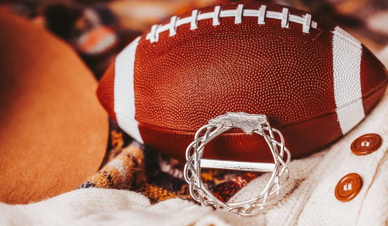 May include: A brown and white football with a silver charm in the shape of the state of North Carolina. The charm is a wreath with the state outline in the center.