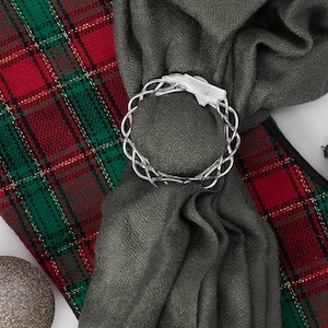 May include: A silver Celtic knot ring ornament is used to hold a gray scarf on a red and green plaid fabric. The scarf is tied in a knot and the ornament is placed on top of the knot. There are also silver and black Christmas ornaments in the image.