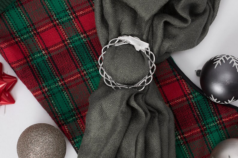 May include: A silver Celtic knot ring ornament is used to hold a gray scarf on a red and green plaid fabric. The scarf is tied in a knot and the ornament is placed on top of the knot. There are also silver and black Christmas ornaments in the image.